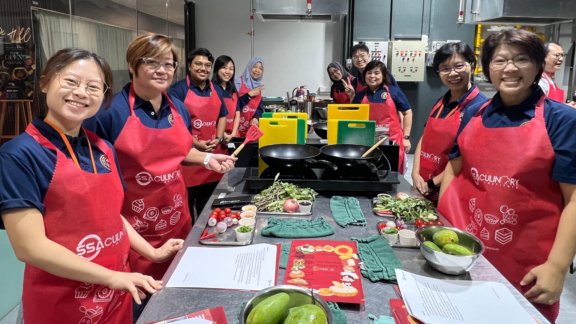 Group of staff in red aprons posing behind a kitchen counter with fresh ingredients and woks.