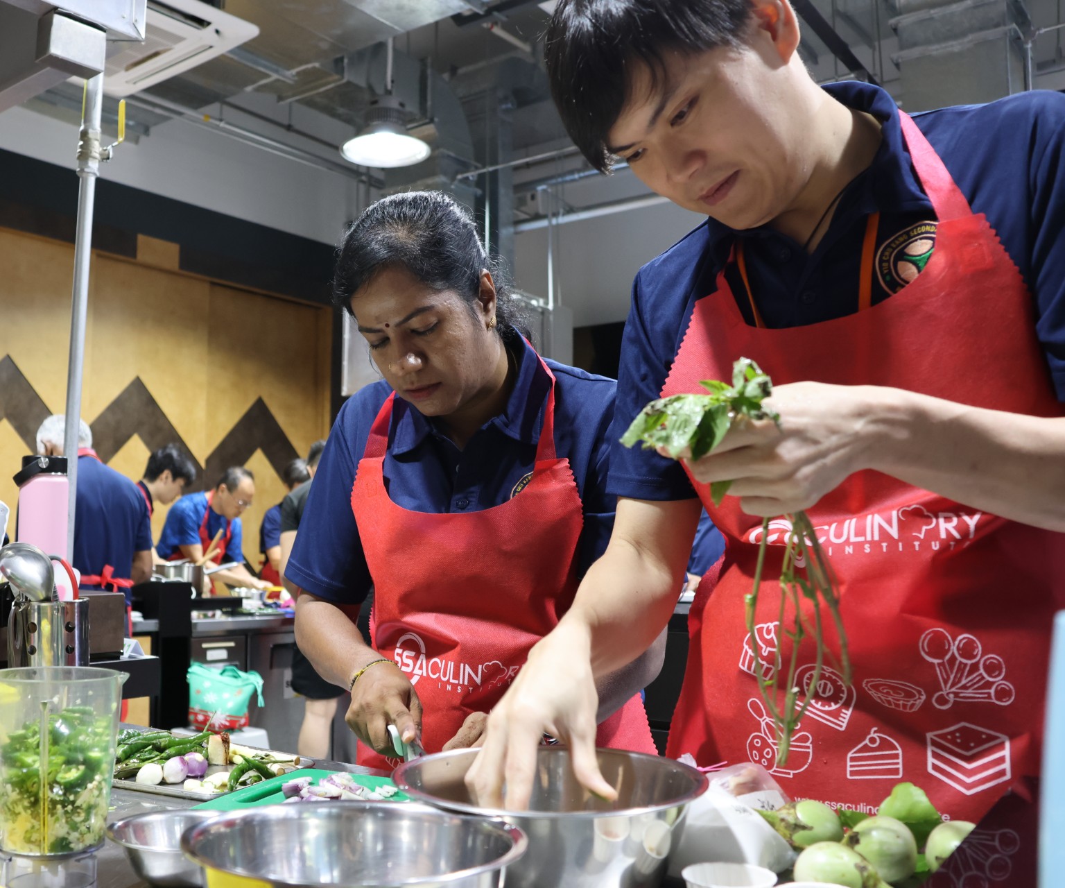 Two staff members in red aprons carefully chopping vegetables and prepping herbs in a kitchen.