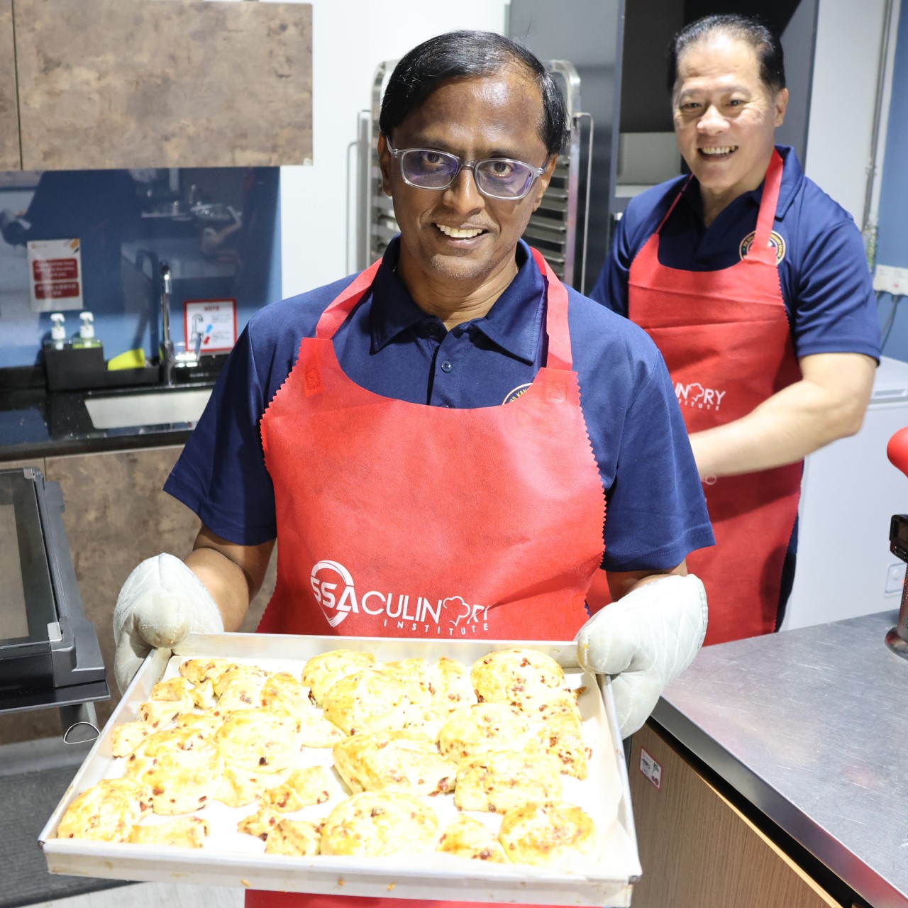 A man in a red apron and oven mitts proudly holding a tray of freshly baked golden pastries.
